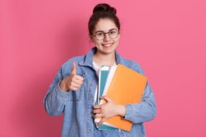 Excited young student wearing denim jacket and eyeglasses, holding colorful folders and showing thumb up, looking directly at camera, dark haired female with hair bun expressing positive emotions.