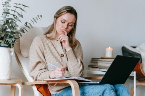 Thoughtful woman taking notes in planner while working with laptop